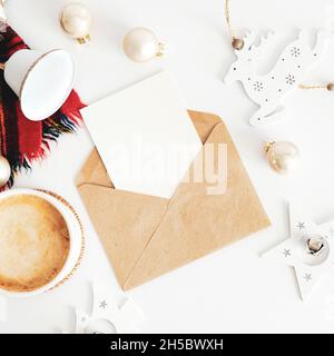 Weihnachtskomposition. Bastelpapier-Umschlag mit leerem Papierkartenmockup, Schal, Kaffeetasse, Vintage-Weihnachtsdekoration auf weißem Tisch. Flach legen, bis Stockfoto