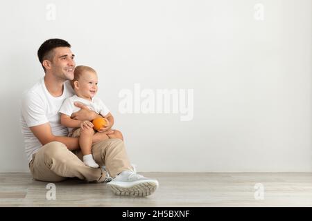 Junger Vater Sitzt Auf Dem Boden Mit Kleinkind Sohn, Schaut Weg Und Lächelt Stockfoto