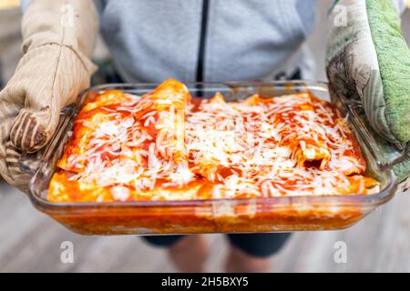 Person hält Backblech Glasschale mit hausgemachten mexikanischen Essen Enchiladas mit Tomatensauce, Tortilla und geschmolzenem Käse Stockfoto