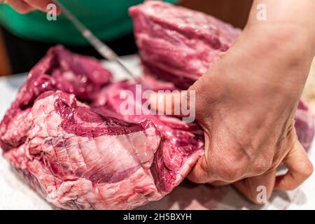 Rotes, rohes Rindfleisch oder Lammfleisch, ganzes Bein, mit einer Frau, die Messerstücke auf dem Schneidebrett zum Kochen verwendet Stockfoto