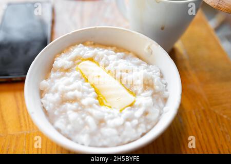 Reisbrei mit Milch und Gras gefüttert gelbe Milchbutter in weißer Schüssel auf rustikalem Holzküchentisch mit Tasse Kaffee und Handy-Smartphone Stockfoto