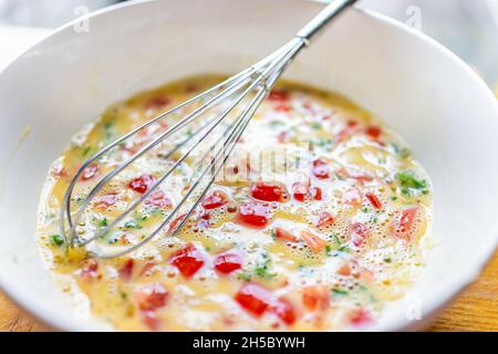 Makro Nahaufnahme der weißen Schüssel Teller mit Ballon Schneebesen Kochen Omelette mit Tomaten, Koriander und roten Paprika Gemüse in der hausgemachten Küche Stockfoto