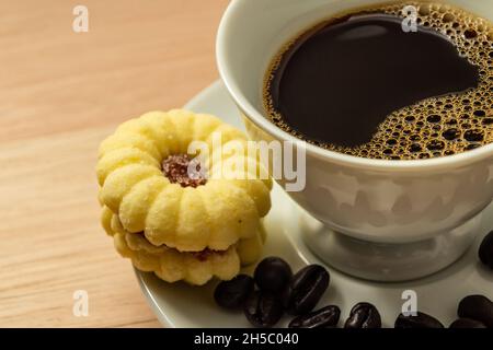 Eine weiße Tasse mit Kaffee und einem Keks und Kaffeebohnen am Rand der Untertasse über einer Holzoberfläche. Stockfoto