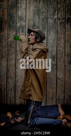 Junkie-Mann mit Mütze grüner Anorak und blauer Jogginghose in der Nähe einer alten hölzernen Wand Stockfoto