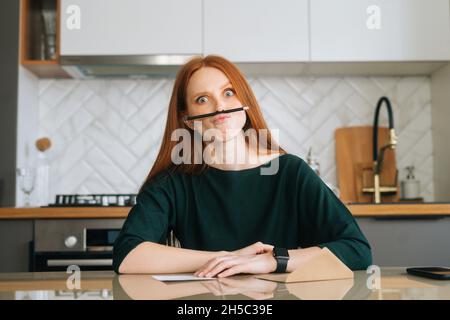 Porträt einer lustigen attraktiven jungen Frau, die Pause macht, einen faulenden Grimace-Bleistift zwischen Nase und Lippen, eine dicke Puppe, die am Tisch sitzt. Stockfoto