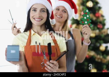Zwei Friseurinnen in Weihnachtsmannmützen halten Scheren und Kämme mit einem Kalender auf dem Hintergrund des Neujahrsbaums Stockfoto