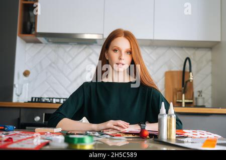 Porträt einer attraktiven Rothaarigen jungen Designerin, die auf einem Tisch mit Set für kreative Arbeit sitzt und die Kamera anschaut. Stockfoto