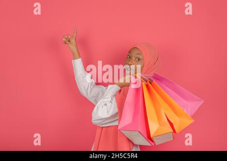 Profil einer aufgeregten jungen muslimischen Frau, die mit den Fingern zeigt und viele helle Einkaufstaschen hält und auf einem rosa Hintergrund auf die Kamera blickt. Stockfoto