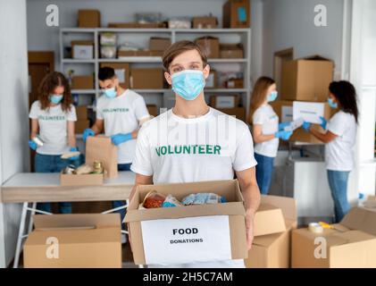 Wohltätigkeitszentrum. Junge männliche Freiwillige in medizinischer Maske, halten Box mit Konserven und Getreide, packen Spenden im Büro Stockfoto