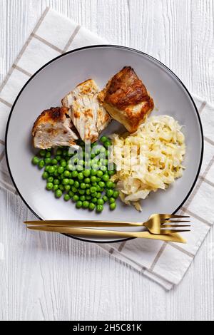 Gebratenes Schweinelende und Sauerkraut auf einem Teller mit grünen Erbsen auf einem weißen Holztisch mit Besteck, Draufsicht, Nahaufnahme Stockfoto