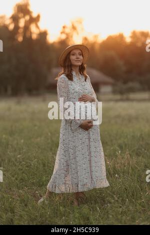 Eine Schwangere im Hintergrund des Sonnenuntergangs posiert und hält ihren Bauch mit den Händen am Abend in der Nähe der Farm auf der Wiese. Eine glückliche Schwangerschaft. Stockfoto