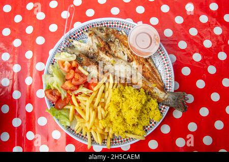 Ein Teller mit gegrilltem Fisch und Pommes auf einer leuchtend roten und weißen Tischdecke auf der Golborne Road in West London, England. Foto: SMP NEWS Stockfoto