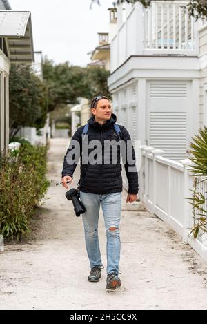 Glücklicher junger Mann Fotograf mit Kamera zu Fuß in Florida Panhandle Winterstadt in Mantel mit weißen neuen Urbanismus Architektur Häuser Stockfoto