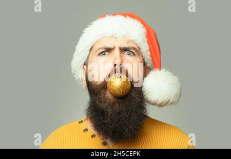 Verrückter Mann mit langem Bart und Schnurrbart auf ernstem Gesicht mit Weihnachtskugel im Mund auf grauem Hintergrund. Nahaufnahme lustige Gesicht Porträt Stockfoto