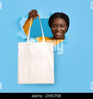 Junge schwarze Frau hält Öko-Tragetasche mit Mockup für Ihr Markenlogo-Design durch Loch in zerrissenem blauem Papier Stockfoto