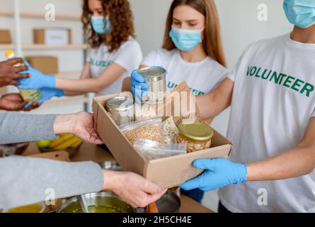 Junge Freiwillige in Masken, die Lebensmittel in Dosen, Gläsern und Verpackungen als Spende für arme Menschen in Kartons verpacken Stockfoto