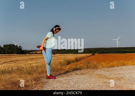 Schöne junge Frau mittleren Alters, die sich im Freien auf dem Feld ausdehnt, mit Musik-Kopfhörern und Windmühlen am Horizont Stockfoto