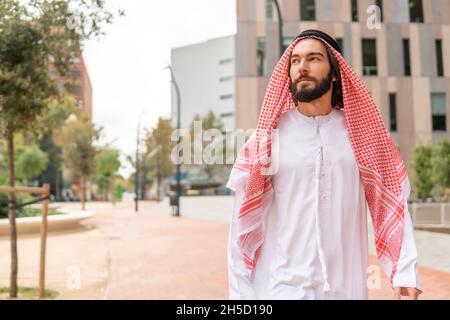Bärtiger arabischer Mann in traditioneller Kleidung, der beim Spazierengehen im Park in der Innenstadt wegschaut Stockfoto