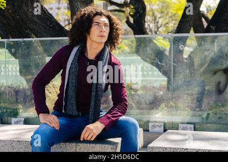 Ein Porträt eines ernsthaften, lockigen jungen Latinos, der Jean-Hosen und ein Hemd trägt und auf einer Betonbank unter einem Baum sitzt. Stockfoto