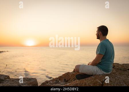 Ein Mann genießt den Blick auf den Sonnenuntergang auf dem Meer, sitzt auf einem Felsen. Seitenansicht, Silhouette. Stockfoto