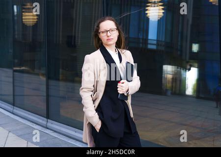 Eine Geschäftsfrau aus dem Kaukasus in einem Mantel und Anzug mit nachdenklicher Optik steht in einer Brille in der Nähe des Geschäftszentrums. Das Konzept der Lösung komplexer Geschäftsprobleme Stockfoto