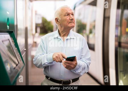 Älterer Mann mit Smartphone, während er auf der Straßenbahnhaltestelle steht Stockfoto