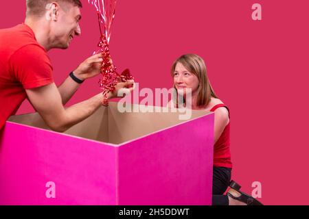 Glücklicher Mann im roten T-Shirt, der aus der großen pinken Schachtel die kleine Schachtel herausnimmt, die in der Hand hält und ihm einen Heiratsantrag machen will Stockfoto