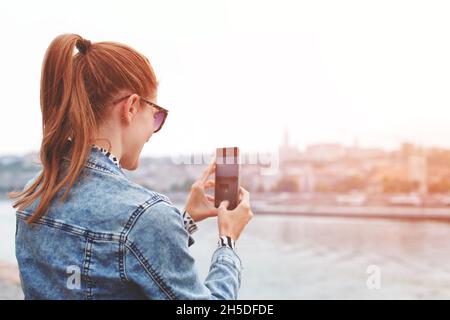 Junge Rotschopf-Frau, die im Sonnenuntergang per Smartphone ein Stadtpanorama fotografiert, Rückansicht, horizontal Stockfoto