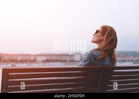 Junge, ausgeglichene, rothaarige kaukasische Frau, die auf einer Bank am Flussufer sitzt und im Sonnenlicht meditiert Stockfoto