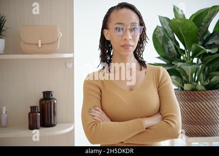 Schöne selbstbewusste schwarze Frau in Brille und Kamelpullover über die Arme und Blick auf die Kamera Stockfoto
