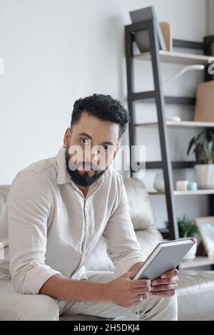 Schöner bärtiger Mann mit Buch oder Planer in den Händen, der auf dem Sofa sitzt und die Kamera anschaut Stockfoto