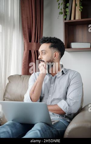 Nachdenklicher Mann mit Laptop sitzt in einem bequemen Sessel und schaut durch das Fenster Stockfoto