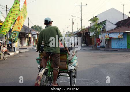 Blick auf den Fahrer und seinen Becak in einer leeren Straße am Morgen mit hellem, klarem Himmel Hintergrund. Ein alter und traditioneller öffentlicher Nahverkehr in Indonesien. Stockfoto