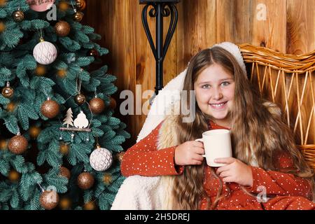 Junges lächelndes langhaariges Mädchen, das auf einer Weidencouch sitzt und einen Becher heißen Getränks in der Nähe des geschmückten Weihnachtsbaums hält. Mädchen an Weihnachten. Stockfoto