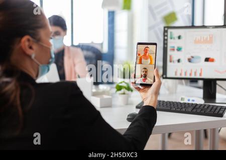 Geschäftsfrau mit Smartphone, die mit Freunden an entfernten Standorten über Online-Videocall-Konferenzen im Büro des Unternehmens spricht. Manager mit Gesichtsmaske gegen Covid19 während der Telefonkonferenz Stockfoto