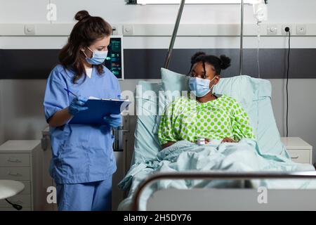 Die Krankenschwester trägt eine schützende Gesichtsmaske, um eine Infektion mit dem Coronavirus zu verhindern, und diskutiert die Behandlung im Gesundheitswesen mit kranken Patienten, die Krankheitssymptome auf der Krankenstation überwachen. Medizinservice Stockfoto