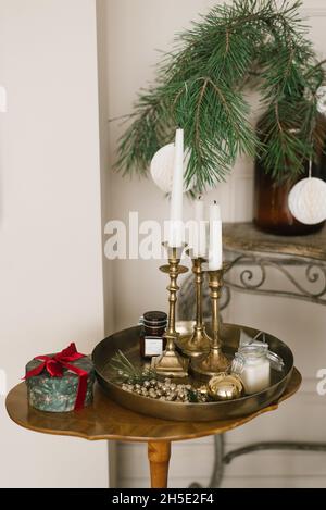 Weiße Kerzen in goldenen Kerzenständern auf einem klassischen runden Tablett, eine Geschenkbox mit einem Bogen auf einem Holztisch. Weihnachtliche Einrichtung im Innenraum Stockfoto