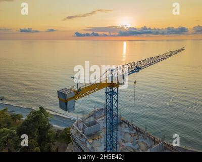 Drohnenansicht eines Turmkrans und eines Gebäudes im Bau vor dem Hintergrund eines fantastischen Sonnenuntergangs an der Küste. Stockfoto