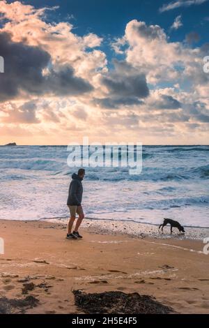 Abendlicht über einem Mann, der mit seinem Hund am Fistral Beach in Newquay in Cornwall entlang der Küste läuft. Stockfoto