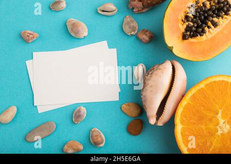 Visitenkarte aus weißem Papier mit reif geschnittener Papaya, Orange, Muscheln auf blauem pastellfarbenem Hintergrund. Draufsicht, flach liegend, Kopierbereich. Tropisch, gesund Stockfoto