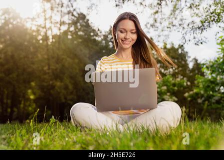 Ganzkörperfoto Frau sitzt auf Gras mit Laptop arbeiten Remote im Sommer Park Stockfoto