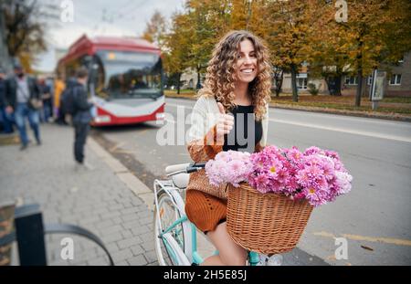 Schöne junge Frau auf dem Fahrrad mit Blumen im Korb sitzen und tun Genehmigung Geste. Fröhliche Frau, die Daumen hoch und lächelt, während sie auf dem Fahrrad auf der Stadtstraße sitzt. Stockfoto