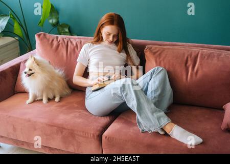 Attraktive rothaarige junge Frau, die Buch liest, sitzt auf einem gemütlichen Sofa mit hübschem Miniatur-spitz-Hund im Wohnzimmer mit moderner heller Innenausstattung. Stockfoto