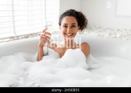 Glückliche junge Frau, die Champagner trinkt, während sie zu Hause in einem heißen Schaumbad neben dem Fenster liegt Stockfoto