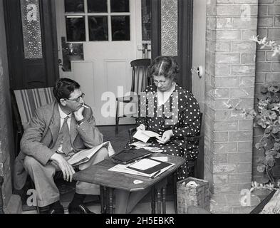 1937, ein historisches Paar, das auf der Veranda vor der Haustür sitzt, mit der Dame an einem kleinen Holztisch, die Papiere in ihrem Portemonnaie aussortierte und dem Herrn auf einem Liegestuhl mit einer Zeitung und einer Zigarette rauchte, England, Großbritannien. Sie tragen die formellen Kleider der Ära, die Dame in einem gepunkteten Kleid und den Herrn in Anzug und Krawatte. Stockfoto