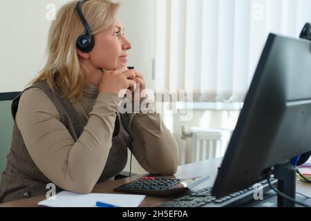 Eine kaukasische Frau mittleren Alters mit einem Headset schaut nachdenklich aus dem Fenster, während sie im Büro sitzt. Stockfoto