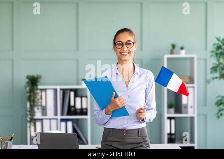 Fremdsprachen-Tutoring. Glücklicher Lehrer mit der Flagge Frankreichs und der Zwischenablage, der neben dem Schreibtisch stand und lächelte Stockfoto