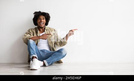 Cooles Angebot. Aufgeregter Schwarzer Mann, Der Auf Dem Boden Sitzt Und Zur Seite Zeigt Stockfoto