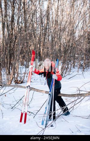 Junge Frau geht durch den Wald. Brunette im Winter Trainingsanzug läuft entlang verschneiten Straße und hält Skistöcke und Skier in ihren Händen Stockfoto