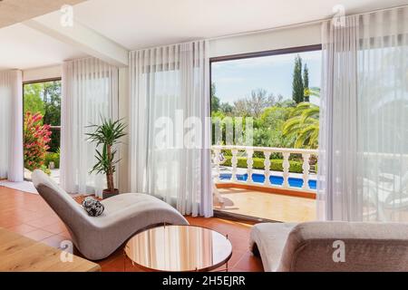 Modernes, traditionelles europäisches Wohnzimmer mit Couchtisch und Blick auf den Pool und den Garten mit Palmen. Für Touristen Stockfoto